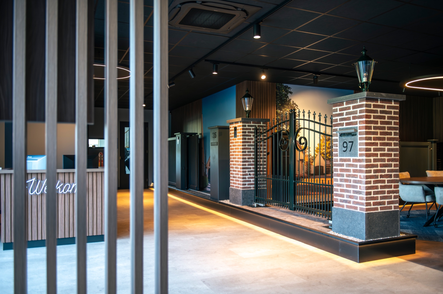 Zicht door houten lamellen op het hekwerkdisplay met bakstenen pijler in de showroom van Van Klompenburg Hekwerken